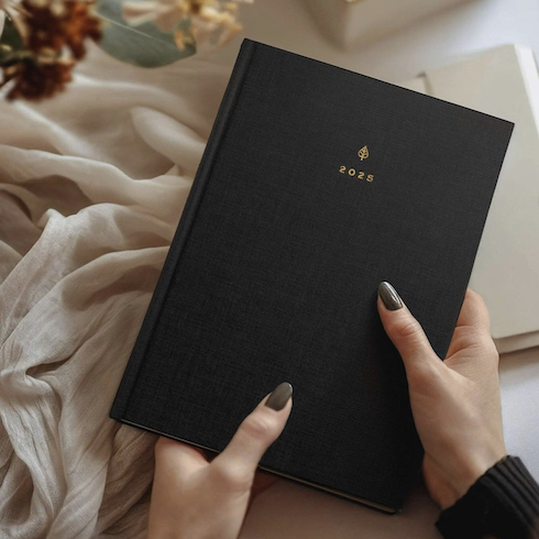It’s a Date Manicured hands hold a new black 2025 Hemlock & Oak planner above a desk decorated with holiday decor as part of HGTV's 10 Gift Ideas for the Women in Your Life