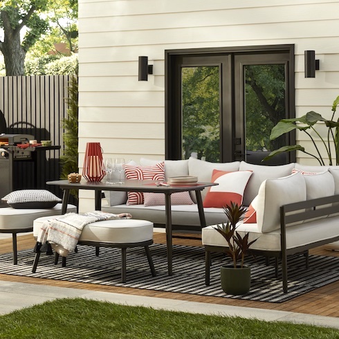 Continuity Supervisor Beautiful backyard wood patio eating area featuring The Home Depot’s Hampton Bay Claxton Steel 5-Piece Patio Sectional Dining Set and Grey Stripe Indoor/Outdoor Area Rug, two potted plants, white and red throw pillows, a woven throw, a white boarded exterior wall with black double doors and two cylindrical exterior lights, a barbeque, and a patch of grass