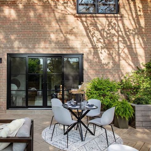 The Earth Laughs in Flowers Outdoor area for al fresco dining with a round dining table, four grey plastic dining chairs, a large sliding black framed door and window set into a tall exterior brick wall with stamped pavers in a herringbone pattern on the ground, potted trees and ferns, and a raised brick garden bed with greenery