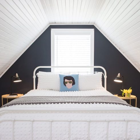 Into Perspective A guest black and white bedroom with a window, white wood panelled walls and ceiling, a white metal bedframe with white bedding, two yellow side tables, two black wall scones, and an Elvis Presley throw pillow