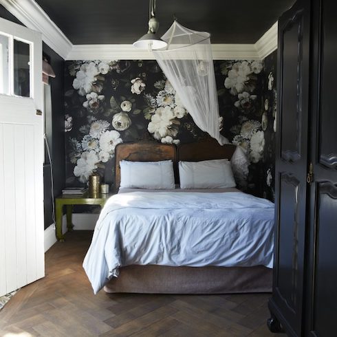Old World, New Money Moody black floral wallpaper in bedroom with dark chevron parquet flooring, a large black armoire, a white mosquito net, a green metal side table, a wood and linen bedframe with white sheets and pillows, a metal pendant lamp, a black ceiling with white molding, and a white exterior door