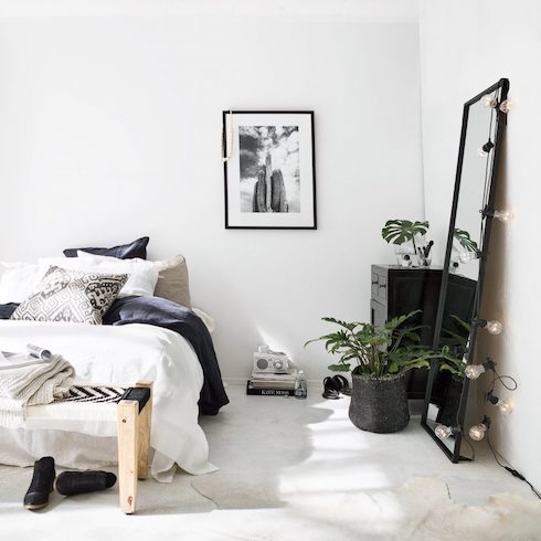 That Loft Life Black and white bedroom in a chic loft with a low bed with white and black linens, a wooden bench with a pair of black boots underneath it, a concrete floor with a white skin rug, a potted plant in a black basket, a black framed mirror with a set of string lights leaning against the white walls, a small black dresser with a small potted palm, a pile of books on the floor and a framed black and white photo on the ground