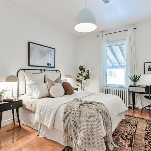 Simply Understated Beautiful staged bedroom in a Toronto house for sale near High Park featuring newly refinished wood floors, white walls, black furniture, black framed artwork, black metal framed bed with white and cream bedding, and a large black and rose coloured Persian rug