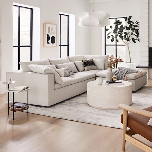 In Harmony Cloud couch dupes featuring a Harmony Modular 3-Piece Ottoman Sectional in a big bright white living room with big black framed windows, light wood floors, modern art on the walls, a large white and grey rug, round coffee table, and a large potted plant in a white pot