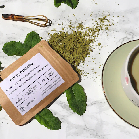 Tea Me Up Packet of Minty Matcha from My Tea Box spills open onto a marble countertop with green mint leaves, a matcha whisk and a green cup and saucer as part of HGTV's 10 Gift Ideas for the Women in Your Life