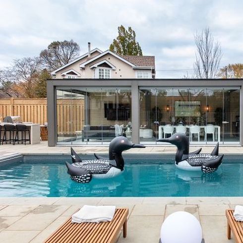 Know When to Fold ’Em A renovated backyard featured on Home Network’s Backyard Builds featuring large sand patio stones, an inground pool with two blow up loon floaties, an eat at bar, and a covered outdoor kitchen with retractable glass walls by Lumon