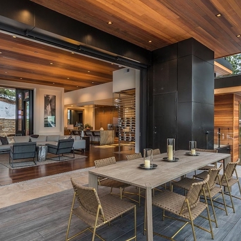 Tile Them Up Home in Aspen, Colorado, with a large retractable wall surrounded by large clack tiles framing an interior with dark wood floors, living room with a white sectional couch and two grey armchairs, wood panelled ceiling with pot lights, a kitchen in the distance with a large marble topped island, and deck with Mbrico Tiles and a large dining table with woven dining chairs