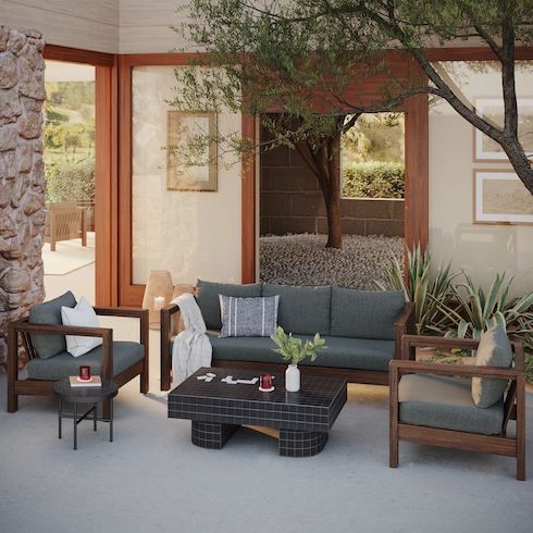 Future Investments Walled outdoor courtyard with Palmera Lounge Chairs and Sofa, Kera black ceramic coffee table, and round side table all by Article, an olive tree, two potted aloes, and a doorway looking out over a rolling hill view