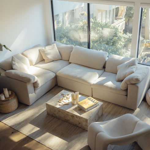 Sky’s the Limit Cloud couch dupes featuring Eternity Modern's EM Sky Sofa modules put together in a living with a wall of windows looking out to a garden below, wood floors, a beige area rug, a curved occasional chair, and stone coffee table piled with books and candles