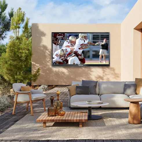 Living on the Outside Sylvox’s waterproof, anti-glare outdoor TV with a football game playing hangs on the wall of an outdoor seating area of an adobe desert house with a sliding wall door, white sectional outdoor couch and armchair, and three outdoor side tables all sitting on a wooden patio next to a cactus and pine tree garden