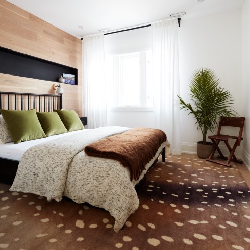 Deer Proof Chic bedroom with a large brown and cream spotted rug, large black framed bed with a brown throw, white sheets and three green pillows, a wood feature wall with a black insert shelf, a large potted palm, a wooden occasional chair, light white drapes and a black curtain rod