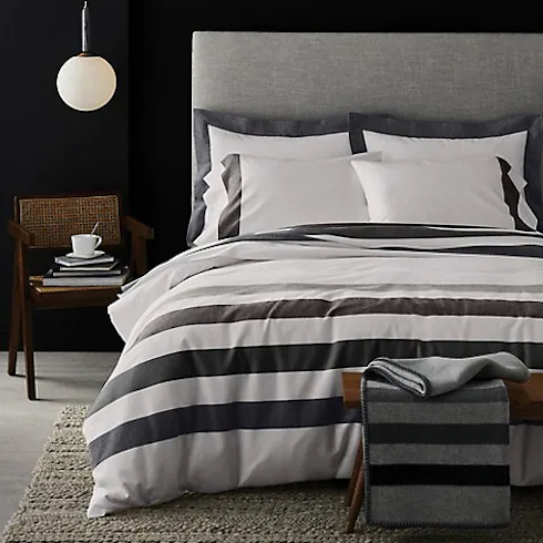 Earn Your Stripes HBC Signature Sterling Stripe Flannel Duvet Cover Set on a large grey upholstered bed in a chic black bedroom with grey floors and a knitted rug, a cane chair with a stack of books and white cup and saucer, and a white pendant lamp hanging from the ceiling
