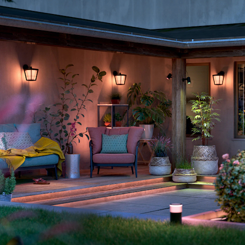 Spotlight on Smart Lights Wall sconces and path lights using Philips Hue Smart Lighting system are seen light up at dusk in a sunken outdoor courtyard, casting soft colourful light over an outdoor sofa and armchair under a covered porch, with multiple potted plants placed around and a grass lawn