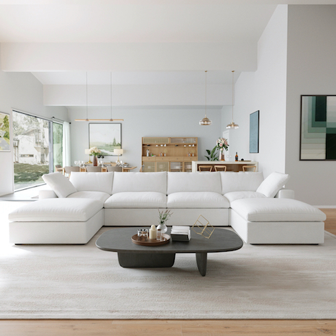 Big Bang for Your Buck Cloud couch dupes featuring Bed Bath & BeyondCloud Modular Sectional Sofa with 2 Storage Ottomans in a large bright living dining area with large glass wall, wood floors, a black coffee table on a beige rug, a dining table set behind along with two sets of pendant lamps, a wooden credenza and art on the walls