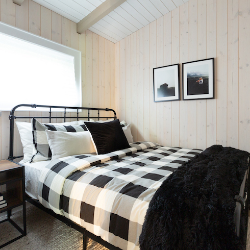 Cottage Core Galore A black and white bedroom in a Muskoka cabin featuring black and white buffalo check sheets, a black faux fur throw, white oak panelled walls, a black metal bedframe, a window, a wood and metal bedside table, and two black and white framed photos on the wall