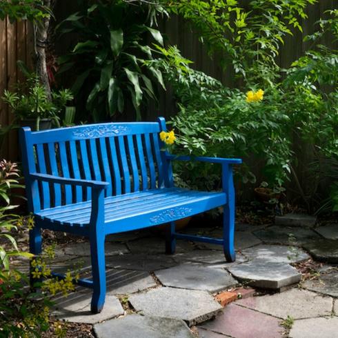 Rest in Peace Blue bench sit on a hexagon tiled ground in a lush garden with palm trees and yellow flowers in a memorial garden