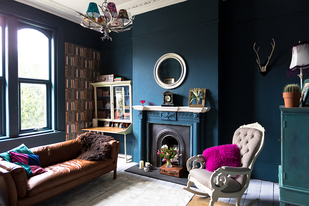 Select a Simple Vase of Flowers Living room with deep blue walls and fireplace. In front of the fireplace is a bouquet of flowers in a vase on top of a vintage suitcase.