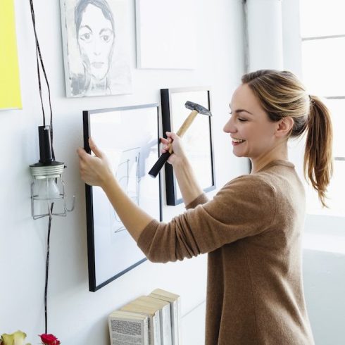 Step Six: Nail It woman holding picture frame and hammer in front of wall