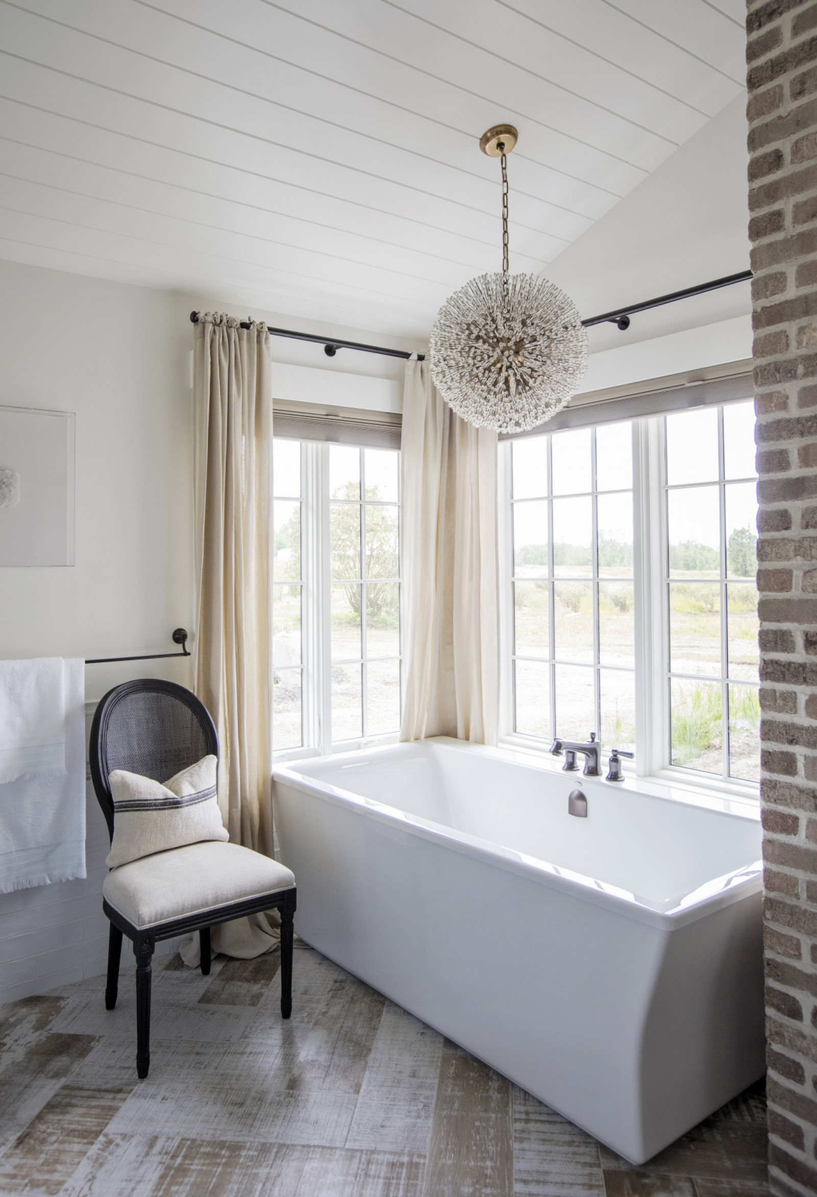 Bath With a View tub in front of window, upholstered chair with dark wood frame, whitewashed chevron floor, orb chandelier