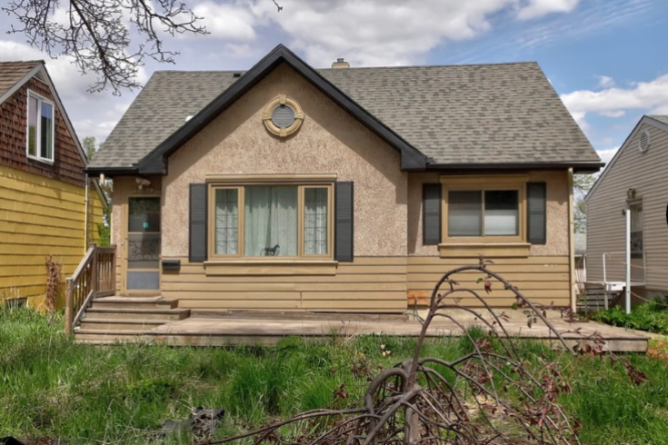 A City Cottage Exterior of a small home with painted stucco, dark grey shutters on two windows, and a main entrance to the side.