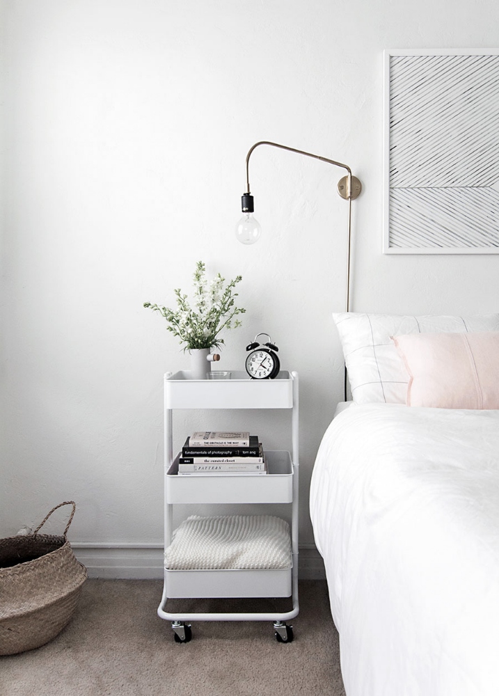 Get Clever With a Cart A white IKEA RASKOG utility cart is used in this bedroom as a three-tier nightstand, housing an extra blanket in the bottom tray, a stack of novels in the middle tray and an alarm clock and vase of fresh flowers in the top tray.