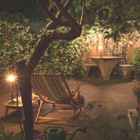 Light the Way Walled garden with a table, bench seat, canvas deck chair and a bicycle at night lit by multiple candles could be part of memorial garden