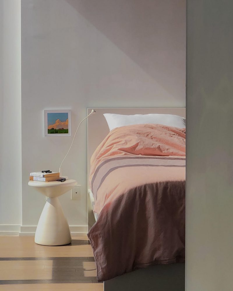 Sculptural Spool A small, sculptural nightstand resembling an hourglass shape holds several books and a lamp next to a bed with pink-tone bedding.