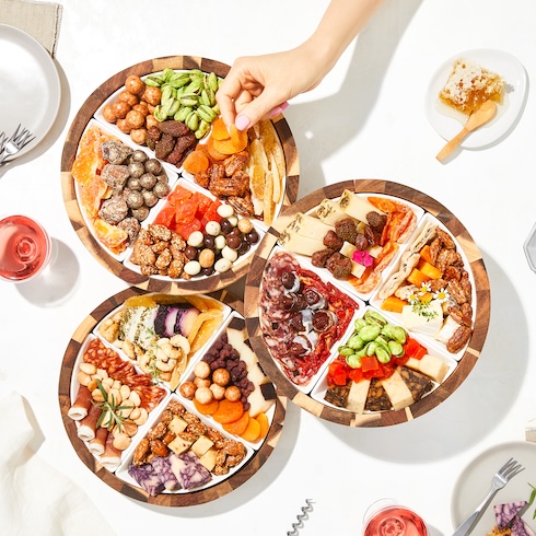Prep Party Food in Advance A hand reaches over three round trays that are each filled with an array of nuts, cheeses, meats, and snacks.