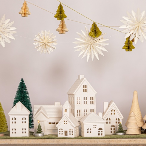 Channel Classic Vibes An array of white Christmas village decorations on table, under hanging paper Christmas trees and stars.