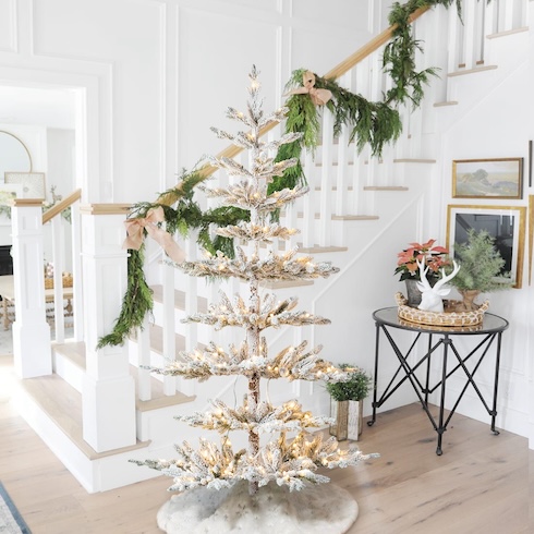 Complement Your Current Decor Style A white-flocked minimalist Christmas tree ists in front of a white staircase that's covered with green garland in a home entryway.