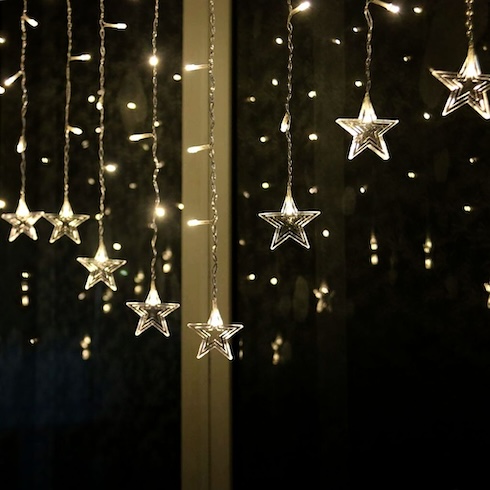 Light’s the Way Many strings of delicate lights with stars at the ends hang in front of a dark window.